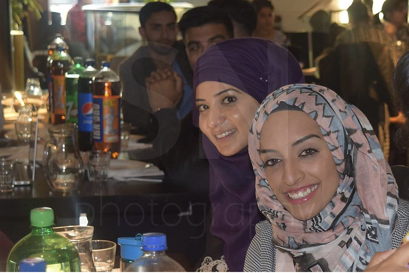 Two women posing and smiling whilst sat a table containing drinks.
